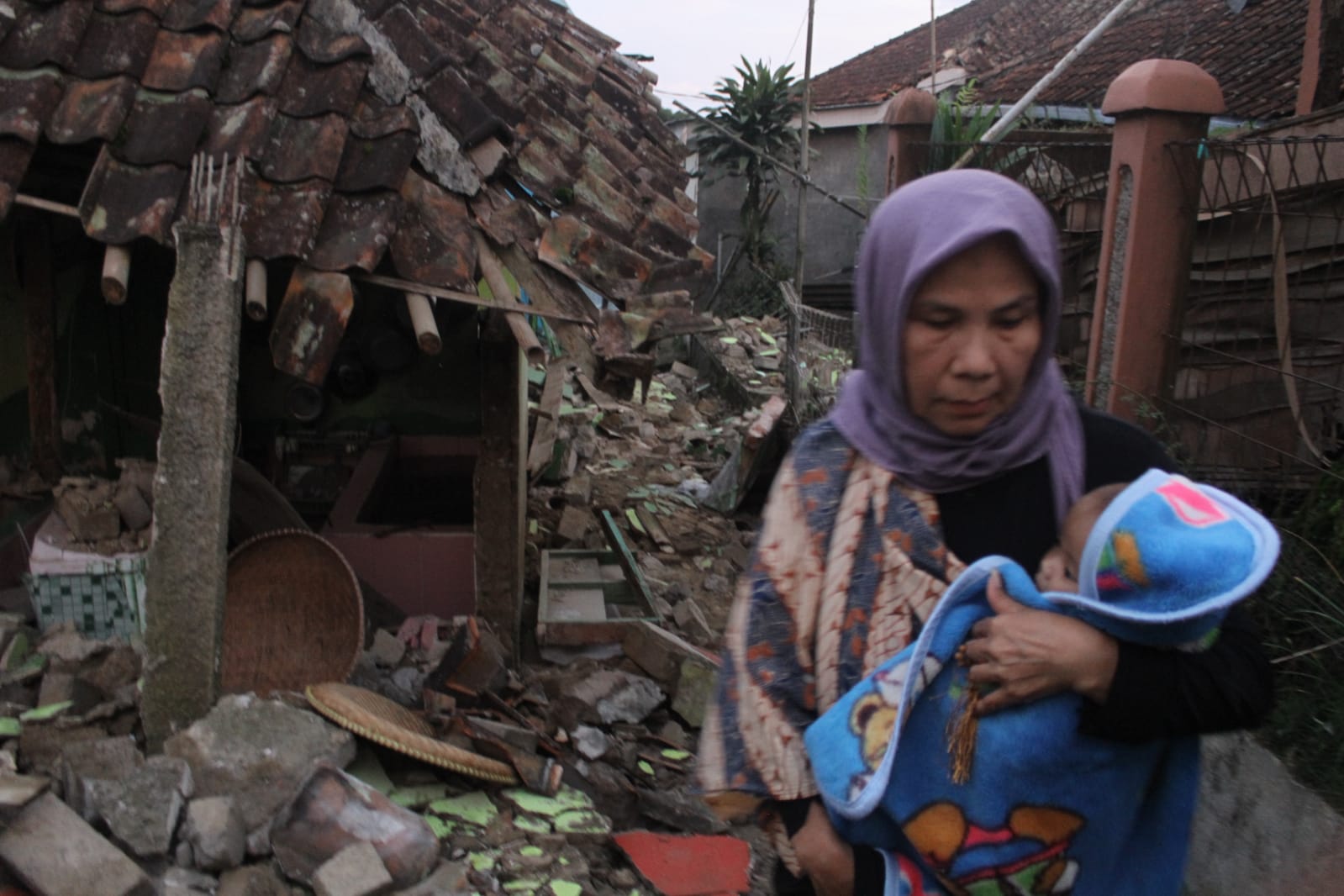 MENYELAMATKAN DIRI: Seorang warga sambil menggendong anaknya melintas runtuhan bangunan yang terdampak gempa bumi Cianjur Senin 21 November 2022. (SANDIKA FADILAH/JABAREKPRES.COM)